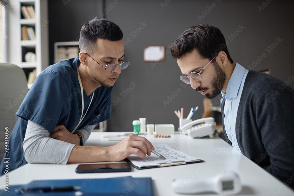Medium shot of Islamic male physician and patient at table looking at medical questionnaire in hospital Medium shot of Islamic male physician and patient at table looking at medical questionnaire in hospital