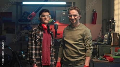 Two mechanics in protective googles in workshop. Femily members spending time together. Father and son standing in home garage, looking at camera and smiling. Maintenance service concept. photo