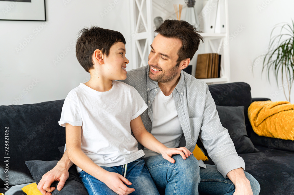 Happy together. Portrait of cheerful father and son sitting on the sofa in cozy living room, looking at each other and laughing, enjoying weekend at home Happy together. Portrait of cheerful father and son sitting on the sofa in cozy living room, looking at each other and laughing, enjoying weekend at home