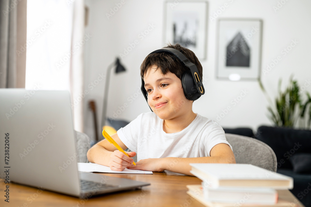 E-studying and homeschooling. Smiling schoolboy studying on the distance from home, using trendy laptop, preteen boy doing homework, writing tasks in the notebook, watching online lessons E-studying and homeschooling. Smiling schoolboy studying on the distance from home, using trendy laptop, preteen boy doing homework, writing tasks in the notebook, watching online lessons