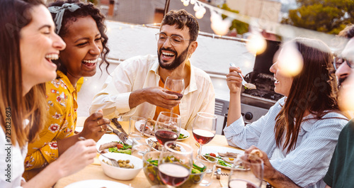 Multiethnic friends having fun at rooftop bbq dinner party - Group of young people diner together sitting at restaurant dining table - Cheerful multiracial teens eating food and drinking wine outside photo