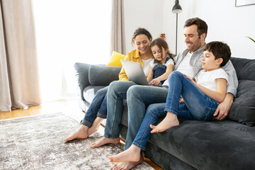 Happy family of four using laptop sitting on the sofa in cozy apartment, parents and two kids talking with relatives, spends time on the weekend together at home