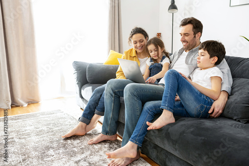 Happy family of four using laptop sitting on the sofa in cozy apartment, parents and two kids talking with relatives, spends time on the weekend together at home