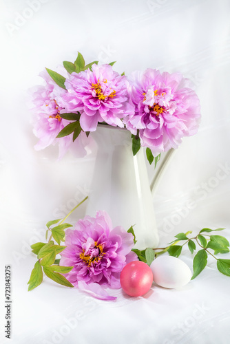 Easter still life with gorgeous pink peonies in a vase and coloured easter eggs on a white background.