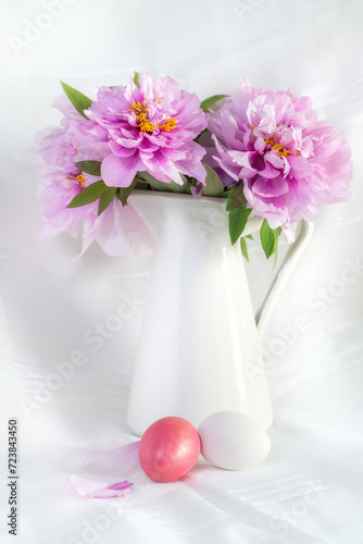 Easter still life with gorgeous pink peonies in a vase and coloured easter eggs on a white background.
