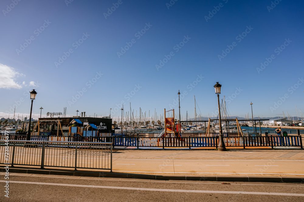Santa Margarita, Spain - January 24, 2024 -  waterfront walkway with palm trees, a marina full of boats, clear skies, and a children's playground in the view.
