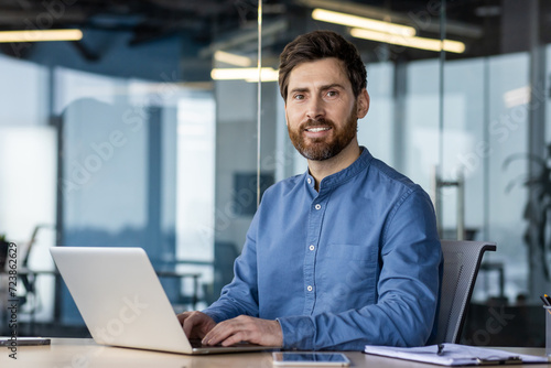 Portrait of a successful and confident young man in a blue shirt sitting in the office at a desk, working on a laptop and smiling at the camera photo