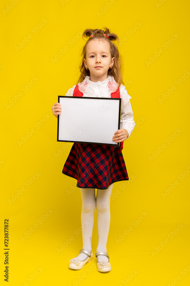 Full body of positive little girl with curly hair in school clothes smiling while standing against yellow background with school backpack and empty paper or banner Full body of positive little girl with curly hair in school clothes smiling while standing against yellow background with school backpack and empty paper or banner