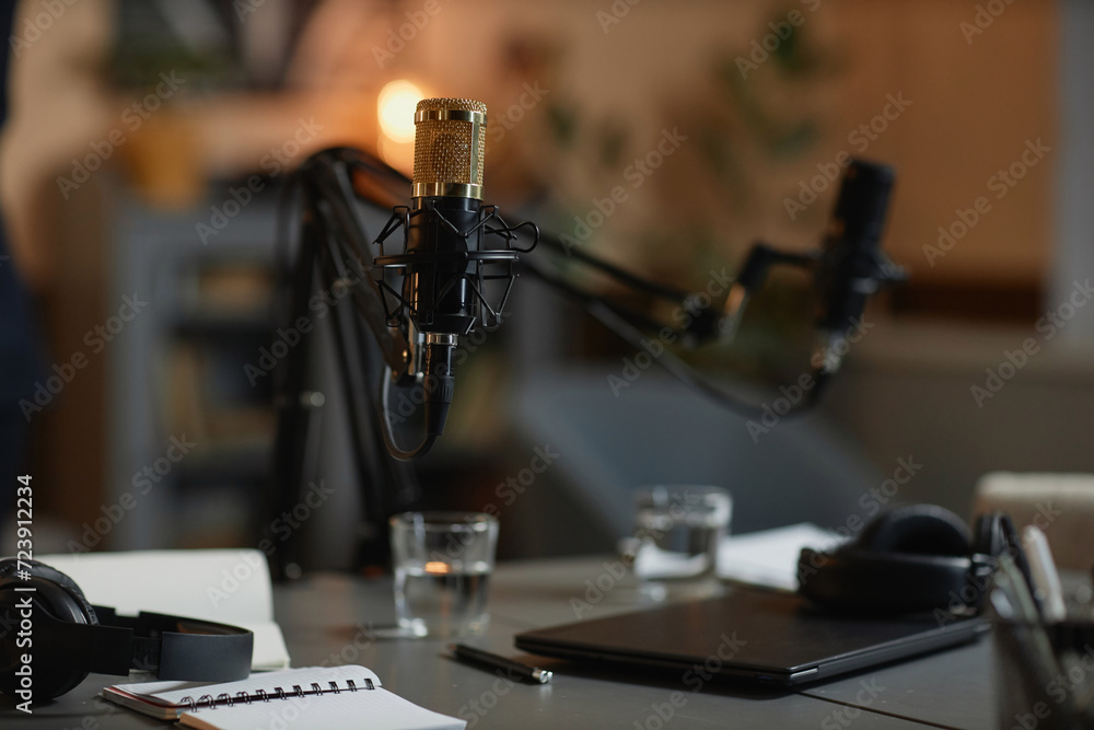 Part of podcast studio with selective focus on gold-colored professional microphone set up on desk with laptop, headphones, notebooks and water glasses