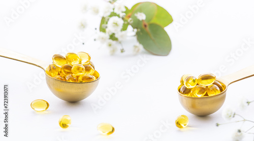 Vitamin D or omega 3 gel capsules in decorative golden spoons on white background decorated with fresh flowers and eucalyptus leaves close up. Health care concept.