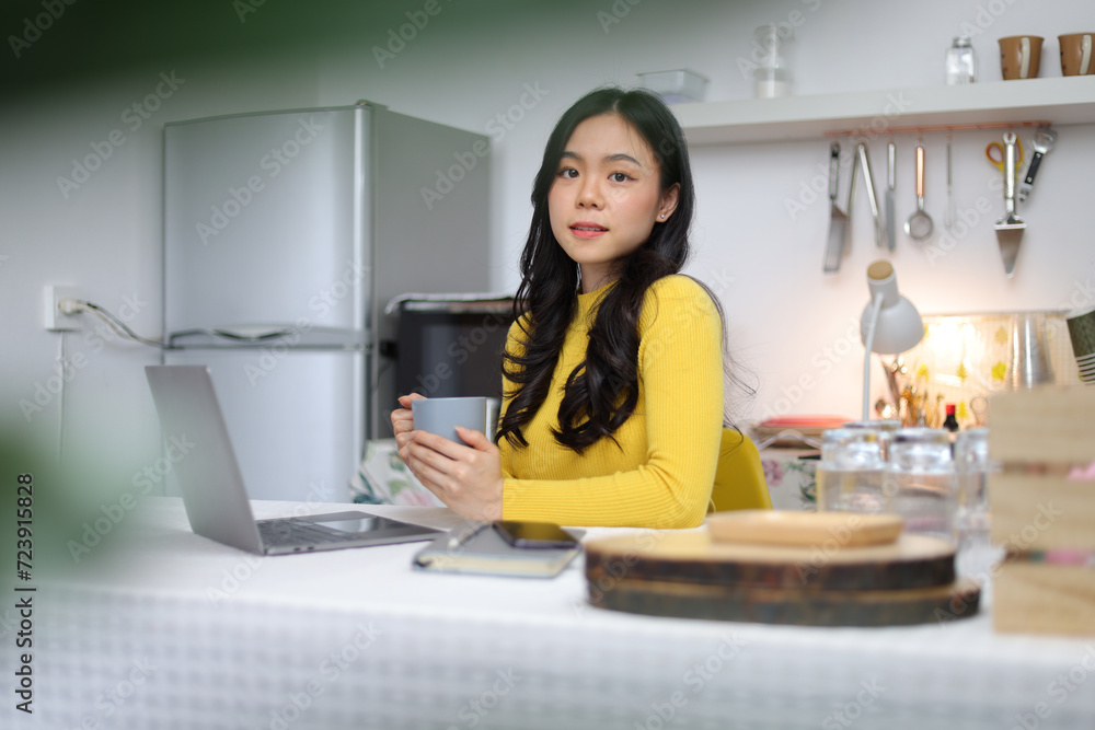 Young Asian woman drinking coffee working with laptop in kitchen at home. Young Asian woman drinking coffee working with laptop in kitchen at home.