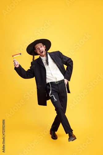 Overjoyed young Jewish man singing and dancing holds groggers in hands against sunny yellow background. Purim, business, festival, holiday, celebration Pesach or Passover, Judaism, religion concept. photo