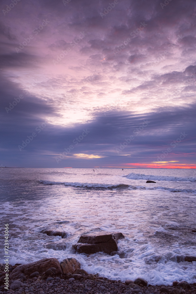 The blue hour over Buzzards Bay in the Atlantic Ocean. Photo taken from the coastline of Dartmouth Massachusetts. Large rocks are in the shallow water and the sky is filled with pink and blue clouds.