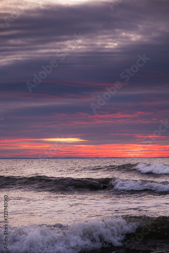 A pattern of three waves making their way to the beach and crashing on the shallow water. Each wave has a whitecap. The ocean is beneath a cloud filled sky lit by the rising sun with pink and orange.