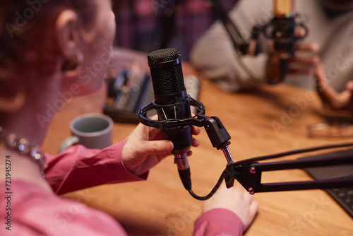 Over the shoulder shot of black condenser microphone and unrecognizable female interviewer arranging stand while talking to podcast guest in studio photo