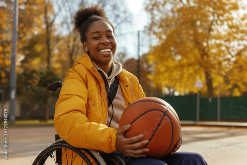 Young beautiful black woman in a wheelchair holding a ball at basketball court. Young disabled basketball player waiting to play on open air ground. Accessibility to sports for disabled athlete. photo