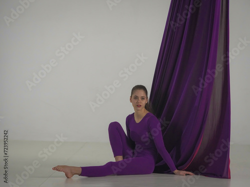 Modern choreography and acrobatics creative advertisement concept. Portrait of female acrobat isolated on white background in studio. Female acrobat sitting on floor next to aerial silk.