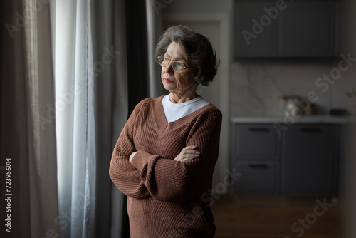 Fotografie Serious and upset senior woman in glasses standing near window with folded arms,