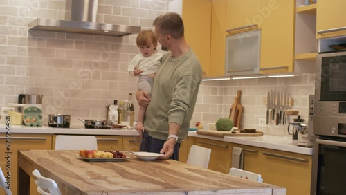 Handheld medium shot of bearded father holding adorable little girl and putting plate with porridge on dinner table in kitchen photo