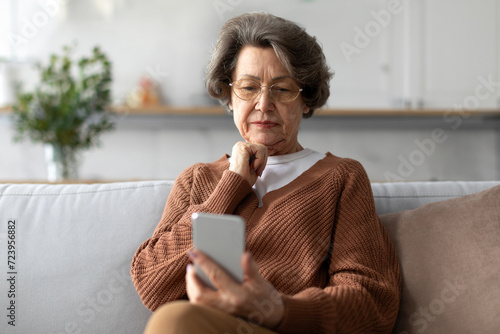 Quadro su tela Worried senior woman sitting on sofa at home and looking at phone screen, readin