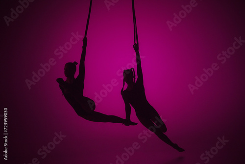 Silhouette of two female acrobats isolated on pink neon background. Girls aerial dancers performing flying element on ropes.