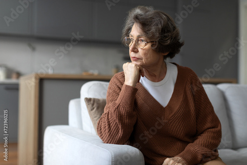 Depressed senior woman sitting on sofa, gazing out of window with thoughtful expression, capturing moment of solitude in her quiet space