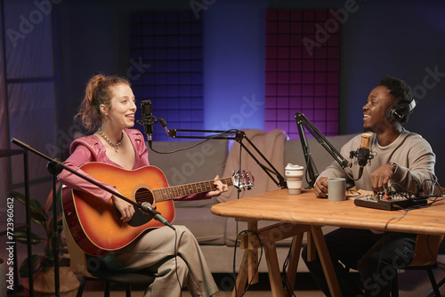 Smiling young girl playing guitar sitting in neon lit podcast studio with African American male host in headphones enhancing sound with audio mixer