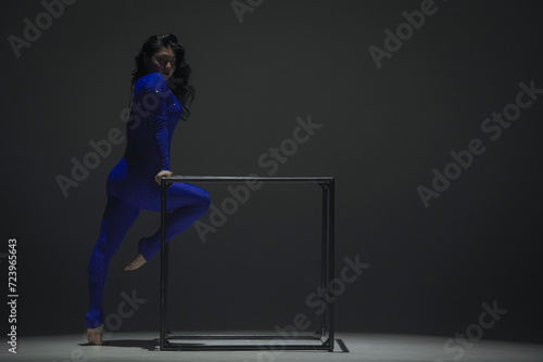 Portrait of female acrobat isolated on black background. Woman gymnastic dancer in blue sparkling bodysuit showing element on a cube.
