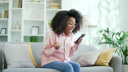 Excited happy young african american female celebrating success by reading great news on smartphone sitting on sofa in living room at home. Smiling black woman satisfied with positive message on phone photo