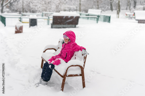 A 7-9 year old girl sits alone on a bench in a snowy park, winter landscape, everything is covered with snow.