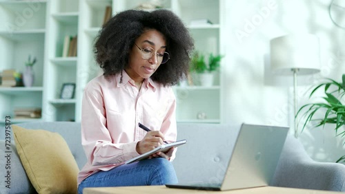 Young african american female student listens to online e-learning, video course using a laptop sitting at home. Black woman at a remote learning seminar or training writes in notebook talks to tutor photo
