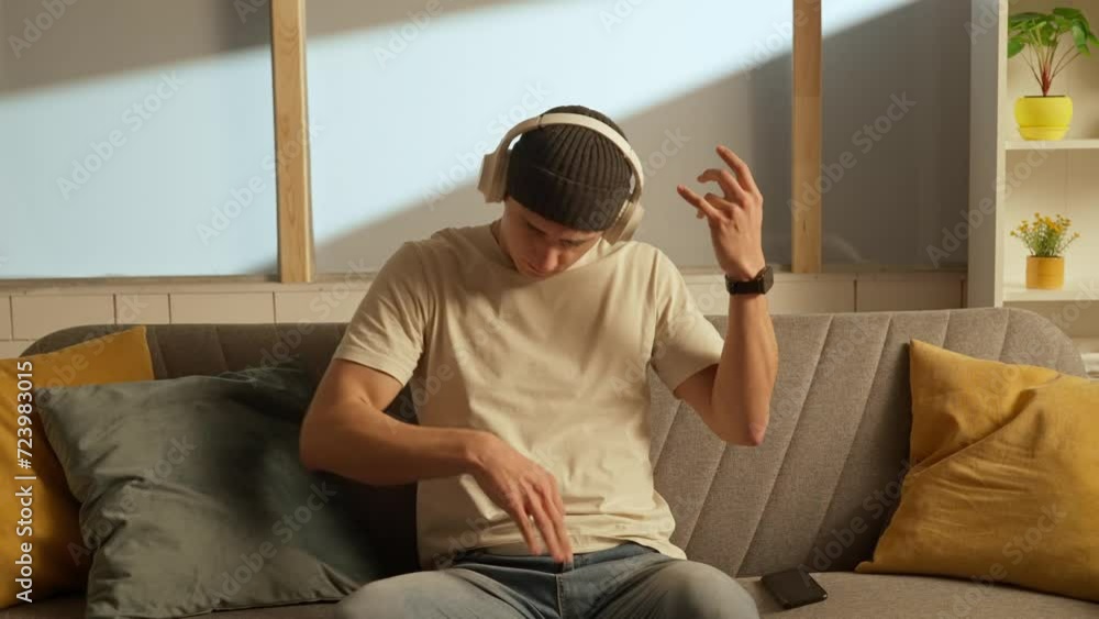 Portrait of young person in the room sitting on the couch. Man in hat on the sofa in headphones listening music playing on air guitar. Portrait of young person in the room sitting on the couch. Man in hat on the sofa in headphones listening music playing on air guitar.
