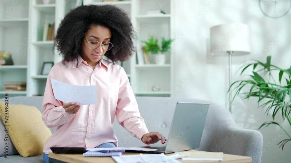Young african american female dealing with household finances using website or app on laptop sitting at home. Black woman holds a utility bill and forms payments online by entering data on a computer Young african american female dealing with household finances using website or app on laptop sitting at home. Black woman holds a utility bill and forms payments online by entering data on a computer