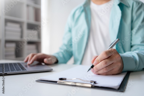 A professional is engaged in writing notes on a clipboard while using a laptop in a modern office setting.