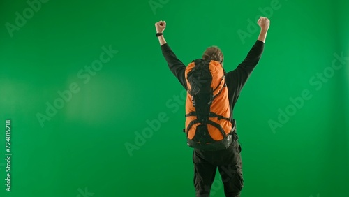 A young tourist joyfully makes a victory gesture, raising his hands in the air. A man with a large backpack on his back stands in the studio on a green screen. Back view.