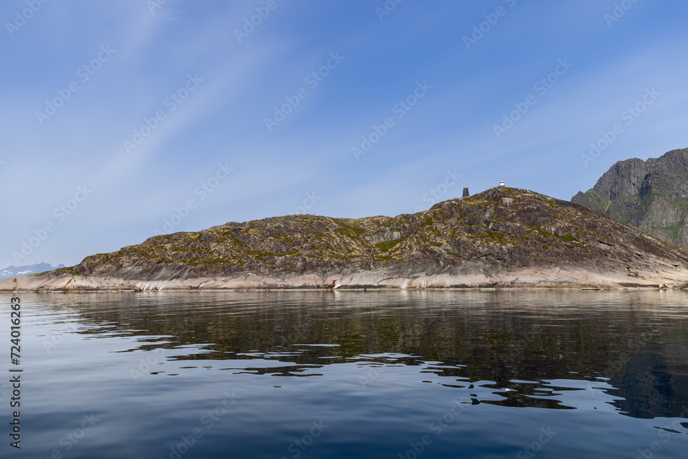 A serene Scandinavian fjord with a lone lighthouse atop a moss-covered rocky island, its reflection on the calm sea water under a clear blue sky. Lofoten, Norway A serene Scandinavian fjord with a lone lighthouse atop a moss-covered rocky island, its reflection on the calm sea water under a clear blue sky. Lofoten, Norway