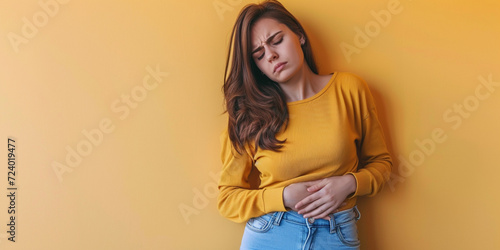 Indoor shot of displeased woman suffering from painful period cramps, holds hands on belly, has pain in stomach, stomachache, upset expression, wears long sleeve shirt on yellow background