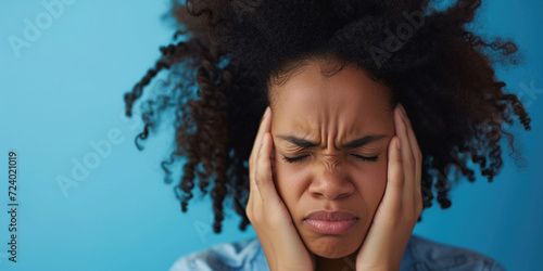 African American young woman holding hands near head while suffering from painful headache migraine on studio blue background. Woman suffer from stress or a headache grimacing in pain photo