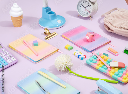 Composition of bright office supplies on table. Colorful notebooks, calculator, pencil case pop it, blue table lamp, erasers, white retro alarm clock.