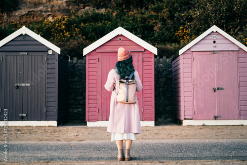 Back view stylish hipster woman with color hair in total pink outfit and backpack looking at wooden beach huts. Off season Travel concept. Seasonal street fashion. Barbiecore style. Simple pleasures photo
