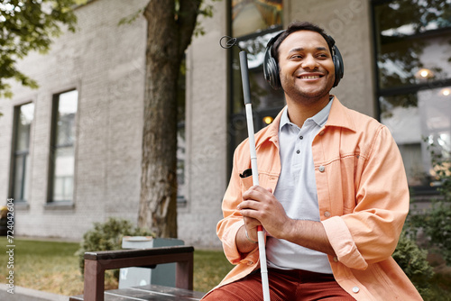jolly indian man with blindness in headphones with walking stick enjoying music while outside photo