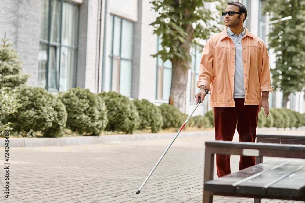handsome indian disabled man with glasses and walking stick in vivid orange jacket walking in park handsome indian disabled man with glasses and walking stick in vivid orange jacket walking in park