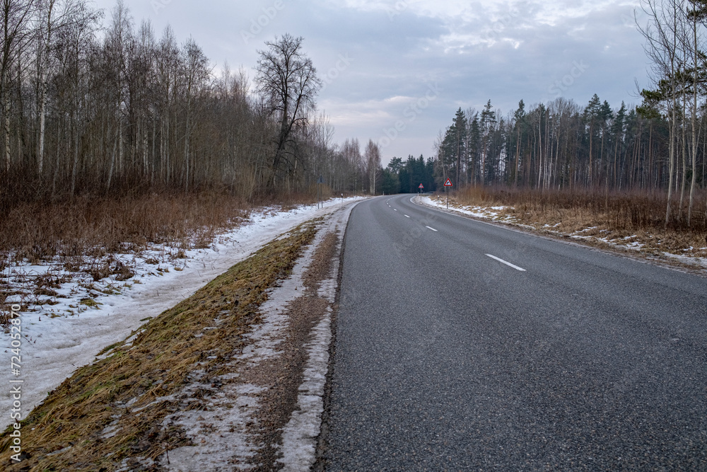 Clean winter road in forest with turns and curve with trees. Winter road with white dividing line at winter season. Melting dirty snow on road side. Clean winter road in forest with turns and curve with trees. Winter road with white dividing line at winter season. Melting dirty snow on road side.