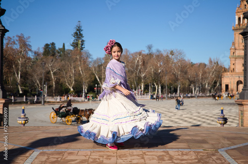 a little girl dancing flamenco dressed in a beige dress with ruffles and purple fringes in a famous square in seville, spain. The girl has flowers on her head and her hair in a bun.