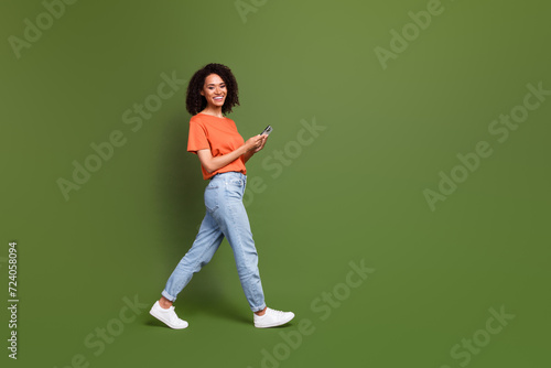 Full length photo of positive cute lady dressed orange t-shirt walking chatting modern device empty space isolated khaki color background © deagreez