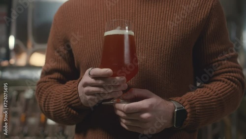 Cropped slowmo of unrecognizable micro-brewery owner wearing knitted red brick color sweater holding glass of fresh bear standing indoors with stainless steel beer production equipment in background photo
