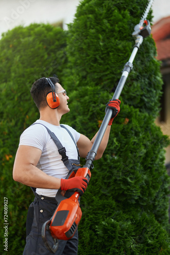 Caucasian handyman in overalls, protective glasses, and gloves pruning hedge on backyard. Professional gardener using electric trimmer for work at garden.