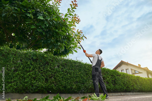Strong caucasian man in uniform and gloves using petrol trimming machine for cutting overgrown trees outdoors. Gardener landscaping during summer time. 