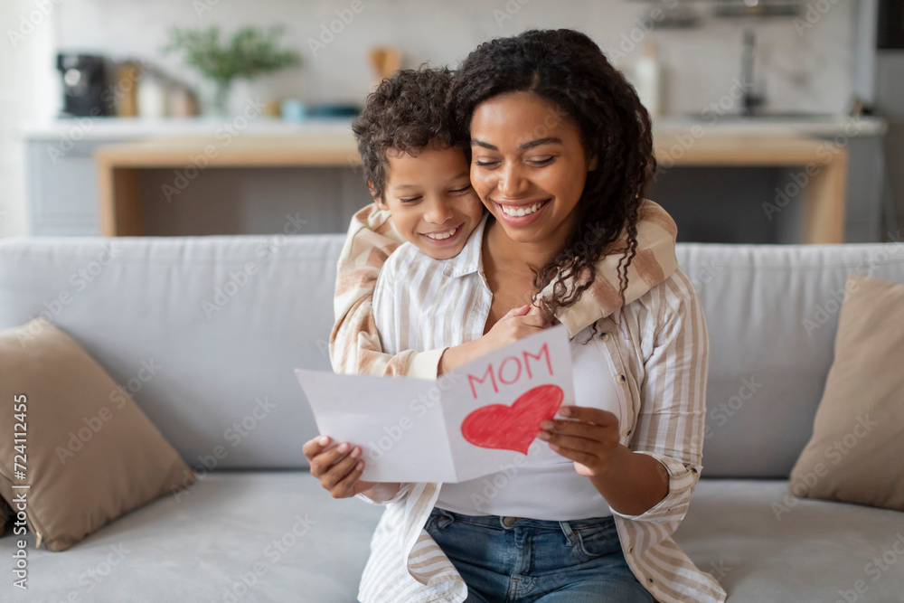 Smiling black mother reading handmade heart card from her cheerful son