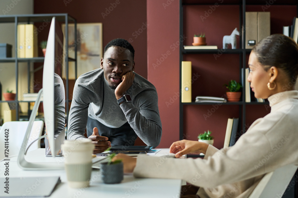Fototapeta premium Young African American businessman bending over workplace of female colleague and looking at her during chat in coworking space